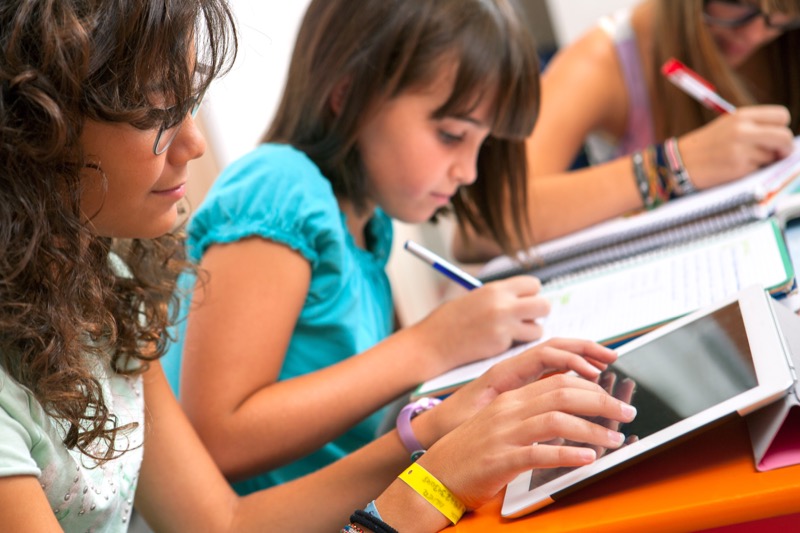 La imagen muestra a 2 niñas, una escribiendo en su cuaderno y otra interactuando con la tablet