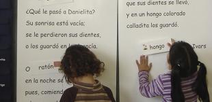 La imagen muestra a dos niñas pegando palabras en un cuento en el pizarrón